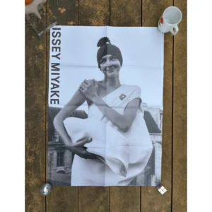 Black and white official brand poster for Issey Miyake Spring Summer 2022 featuring a smiling model with a sculpted headpiece wearing a one-shoulder white dress and clutching a soft white oversized bag.