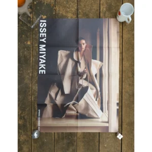 Official brand poster for Issey Miyake Spring Summer 2025 featuring a model seated in a structural paper-like textured beige coat and holding a oversized matching geometric tote bag.