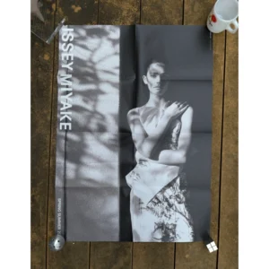 Black and white official brand poster for Issey Miyake Spring Summer 2025 featuring a model in a printed pleated dress standing alongside sunlight and dappled tree shadows cast on a wall