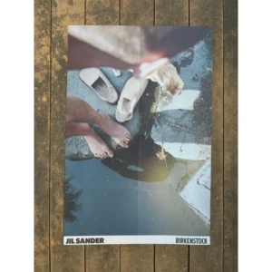 Official campaign poster for Jil Sander Plus x Birkenstock featuring a top-down POV photo of bare feet near a water pond, cream suede clog sandals, and a ginger cat on stone pavement.