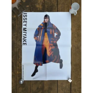 Official brand poster for Issey Miyake Autumn Winter 2022 23 featuring a model in a dynamic pose wearing a long blue coat with orange brushstroke patterns against a clean white background.